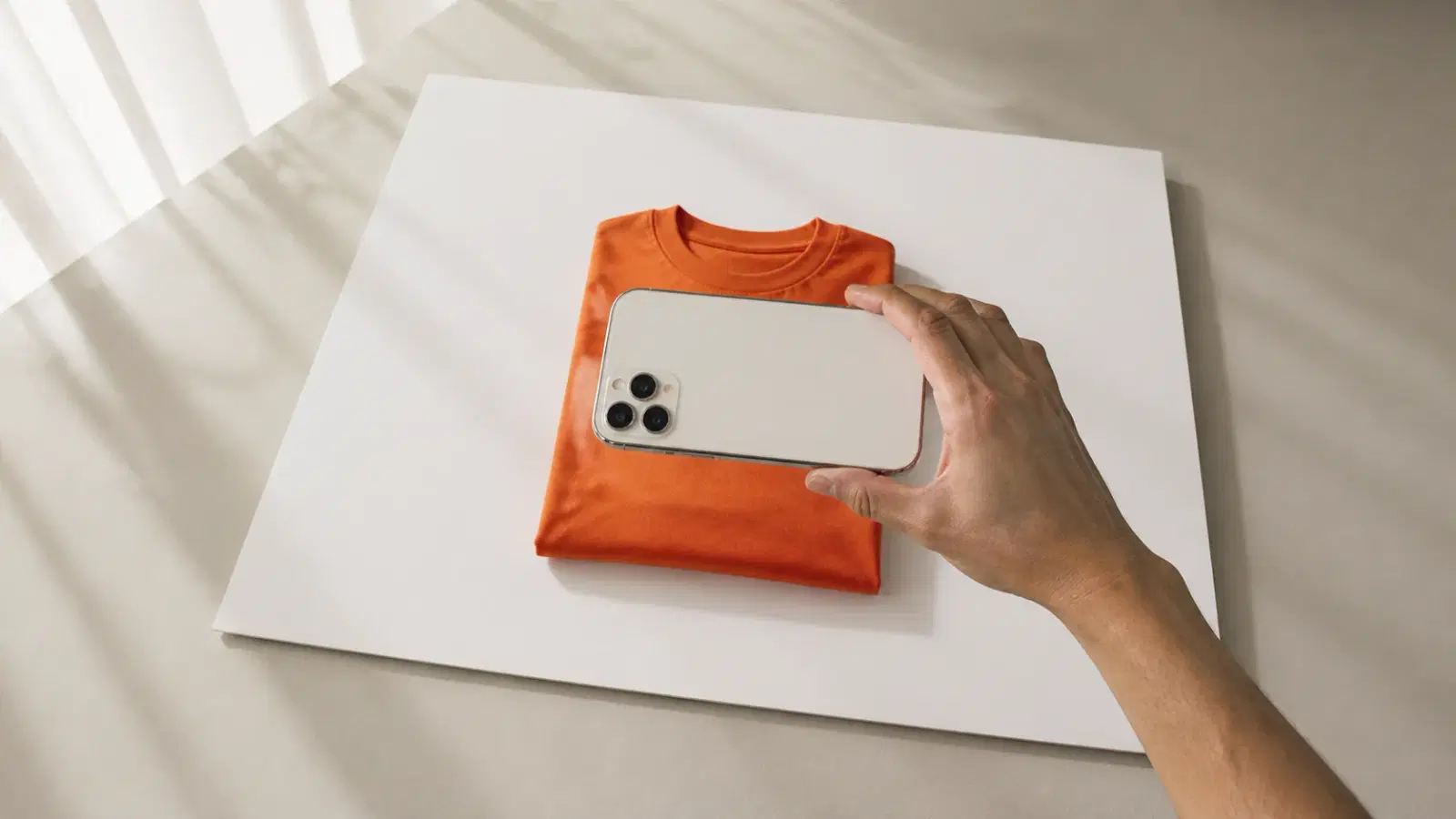 A minimal flat lay clothing photography setup: poster board backdrop, indirect window light, and a phone held parallel to the surface.