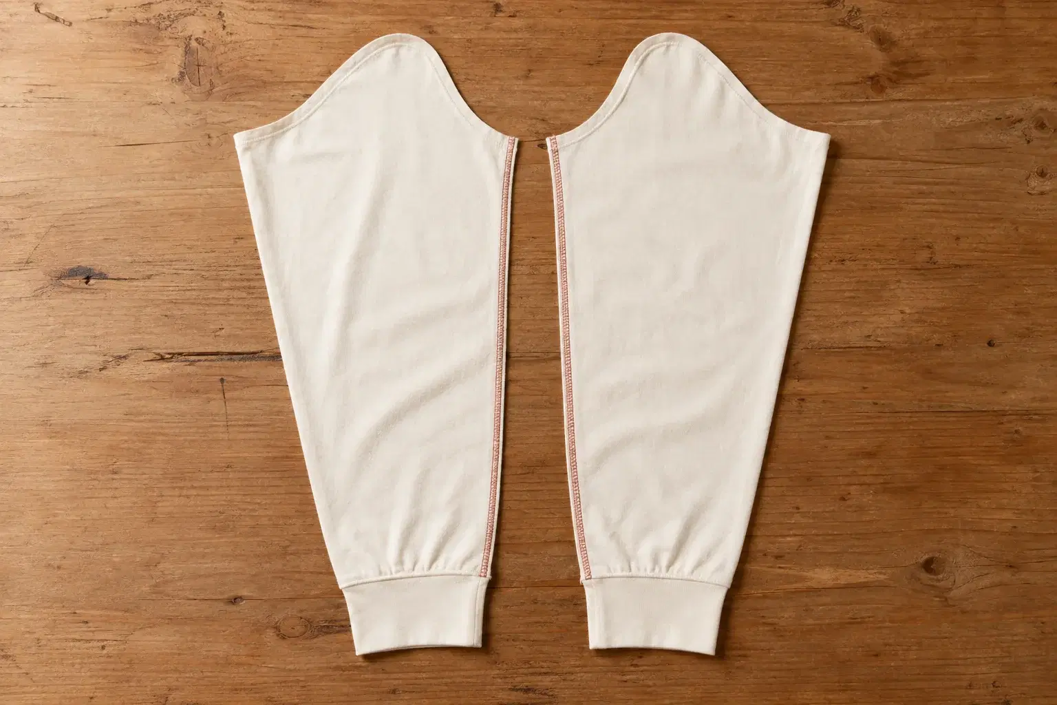 Editorial shot of two blank t-shirt sleeves (left and right) laid side by side on a warm desk as a cut-open pair, showing how mirrored edges must line up at a shared seam line. No text.